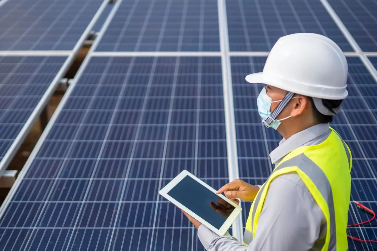 A man in a safety helmet and vest examines a tablet in front of solar panels, illustrating project safety and efficiency in environmental engineering.