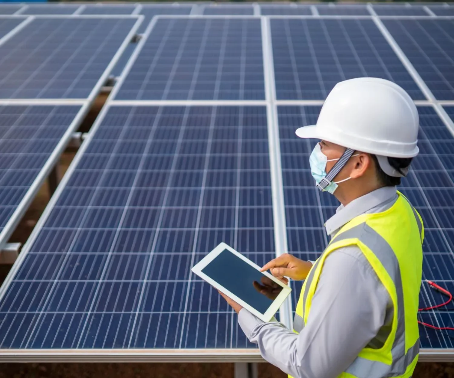 A man in a safety helmet and vest examines a tablet in front of solar panels, illustrating project safety and efficiency in environmental engineering.